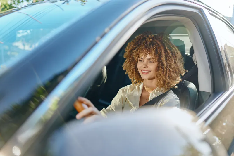 Smiling woman driving car with curly hair in sunny daytime, showing happiness and freedom on a bright urban street.