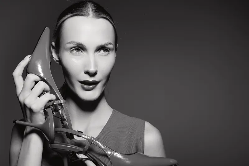 A young Caucasian woman with light skin holds glossy high heel shoes close to her face. Studio portrait with strong lighting and dark background. Concept of fashion and style.
