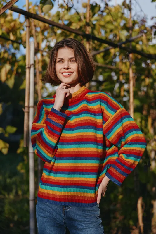 Young woman in early 20s wearing a colorful striped sweater, smiling outdoors by a trellis with vines, casual outfit and knitwear fashion in warm golden hour autumn garden.