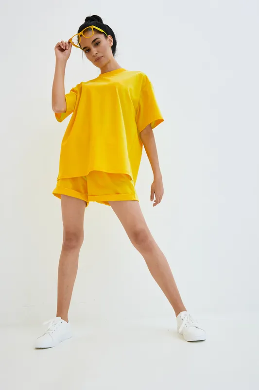 Young woman wearing bright yellow casual outfit and white sneakers poses confidently against plain white background, showcasing modern street style fashion.