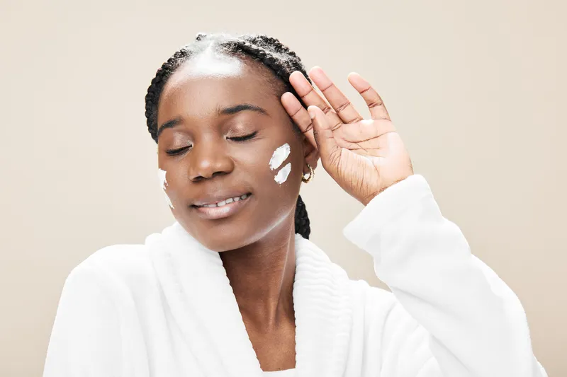 Young Black woman applying skincare product with a peaceful smile in a cozy, neutral toned setting, promoting self care and wellness