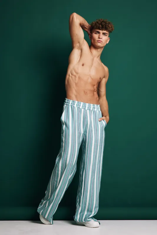 Young man with curly hair standing shirtless against green backdrop with hand behind head. Casual striped pants and confident pose suit fashion editorial and summer style concept for male.