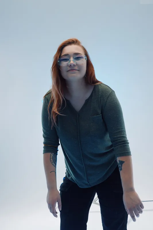 Young woman with red hair and glasses wearing a casual green sweater stands against a light background. Concept of beauty, fashion, and modern style in portrait photography.