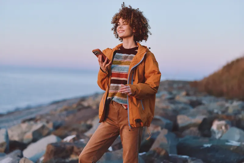 Woman lifestyle outdoors by ocean coast, walking on rocky beach at sunset, wearing an orange jacket, curly hair in sea breeze, calm travel mood with wide horizon and distant waves.