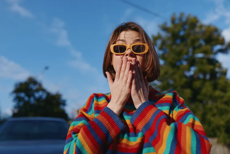 Young woman in her early 20s wearing a rainbow sweater and yellow sunglasses, short hair, surprised expression with hands over mouth outdoors on a sunny urban street.