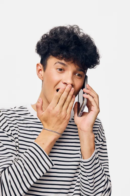 surprised man talking on phone with hand covering mouth, young mixed-race adult, curly hair, casual striped shirt, white background, emotional expression.