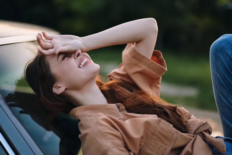 Young woman laughing joyfully while resting on a car hood, dressed in casual clothing, with a vibrant green background, evoking a carefree summer mood.