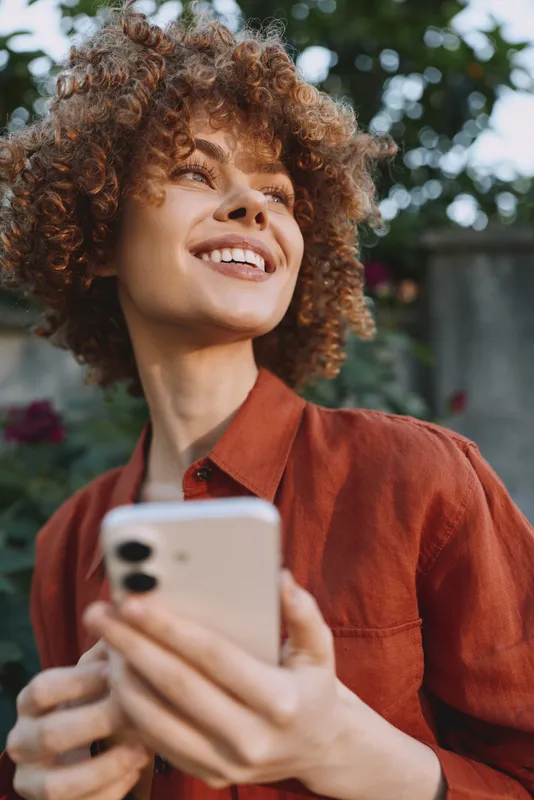 Smiling woman with curly hair holding smartphone outdoors. Casual lifestyle scene with natural light and green background. Focus on joy, connection, and everyday moments