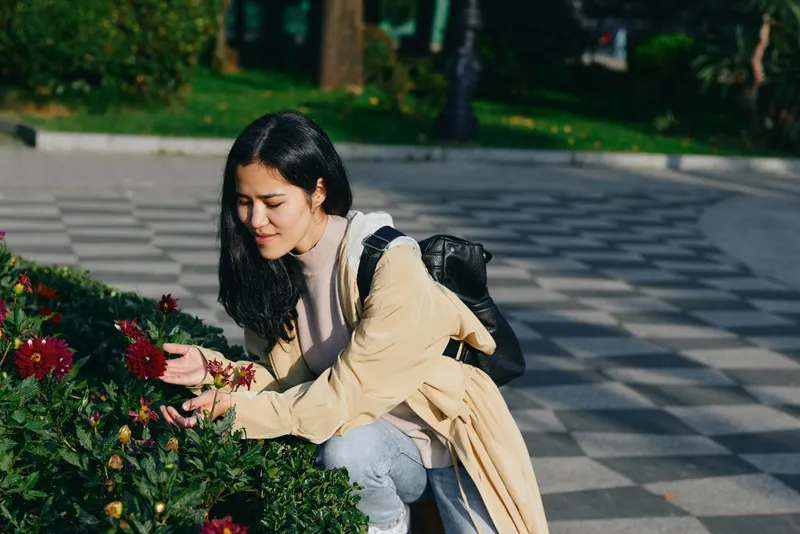 Woman, gardening, flowers, park, nature, outdoor, leisure, sunlight, woman crouches beside vibrant blooms admiring petals in a park garden calm mood casual attire backpack