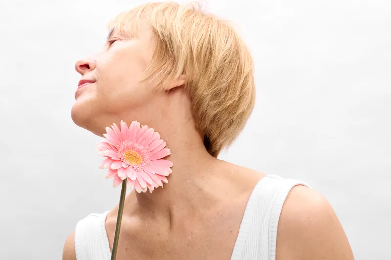 Woman beauty portrait with a pink flower near the neck, serene face, smooth skin, soft light, clean white background, spring mood, calm wellness concept