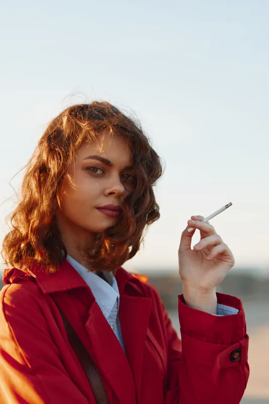 Confident young adult woman in a red coat holds a cigarette outdoors, gazing at the camera with a poised expression, sunlit sky behind, isolated mood.