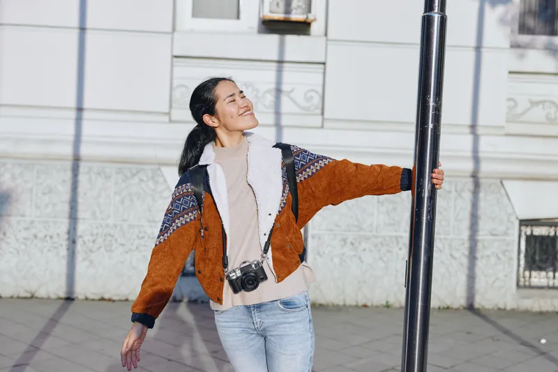 A woman stands outdoors, leaning against a streetlamp in an urban setting, wearing a warm orange and navy jacket, a scarf, and jeans, smiling softly as daylight highlights the scene.