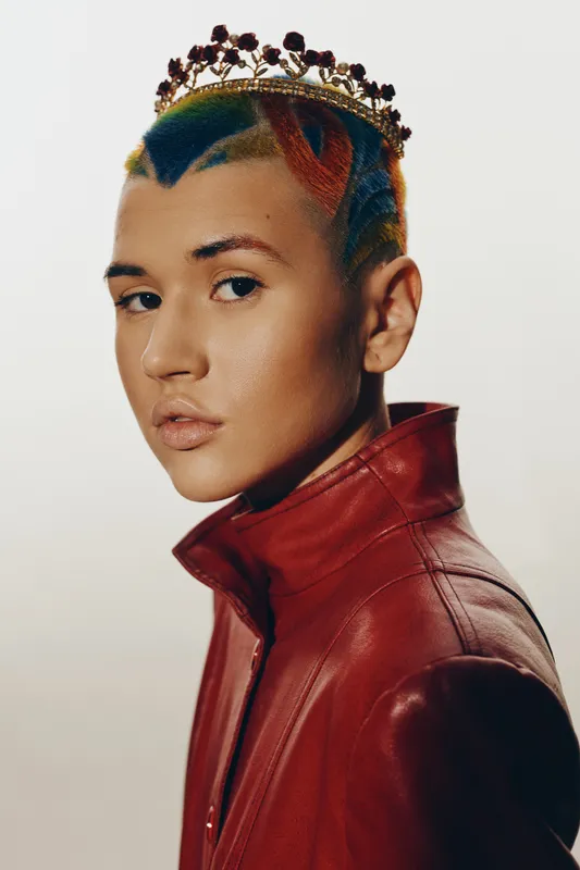 Woman with colorful short hair and crown wearing red leather jacket, showcasing modern fashion and bold style against a neutral background, studio portrait lighting.