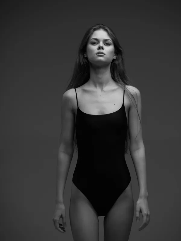 Black and white portrait of a young woman in a black bodysuit with serious expression and long hair against a dark background.