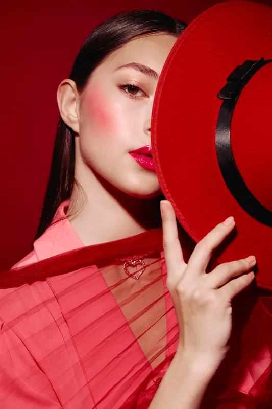 Fashion woman with red lipstick and peach blush hides partially behind a red hat. She wears a pink sheer top, posing in vivid studio light on a red background. Beauty and fashion concept.