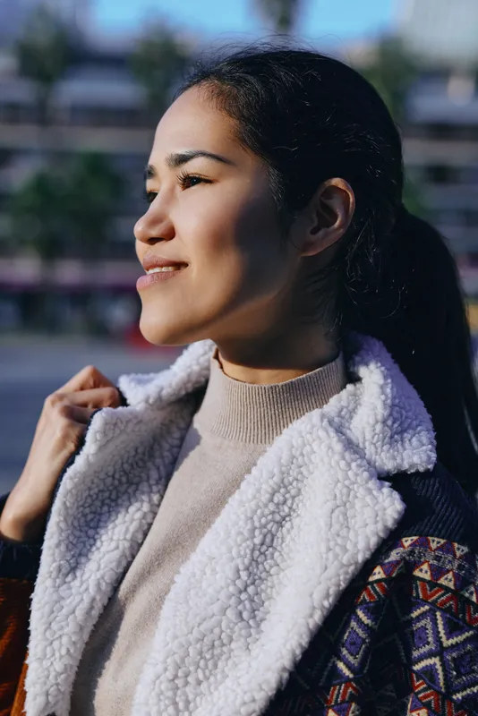 Woman in shearling coat outdoors with sunlight on her face, confident smile, cozy street style, urban backdrop, warm textures, fashionable casual look, candid atmosphere