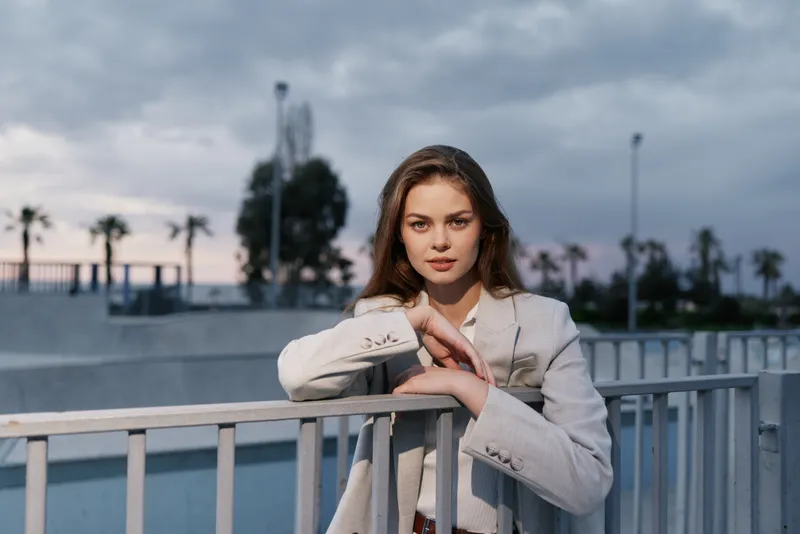 Woman in light suit leaning on metal railing outdoors with cloudy sky and palm trees in background, confident expression, business concept