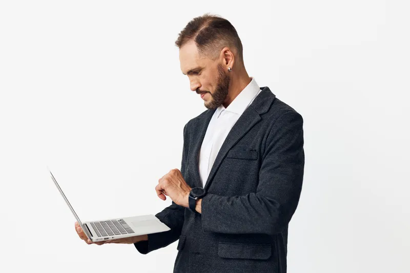 A professional man in business attire stands against an isolated colored background, focused on a laptop, projecting confidence, control, and ready momentum.