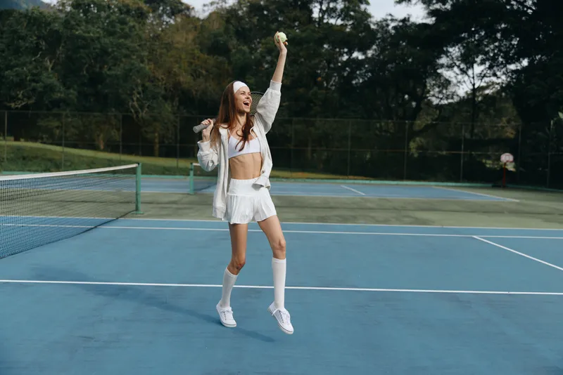 Energetic young woman with long hair celebrates while playing tennis on a sunny day, wearing sporty white athletic wear on a blue court surrounded by greenery