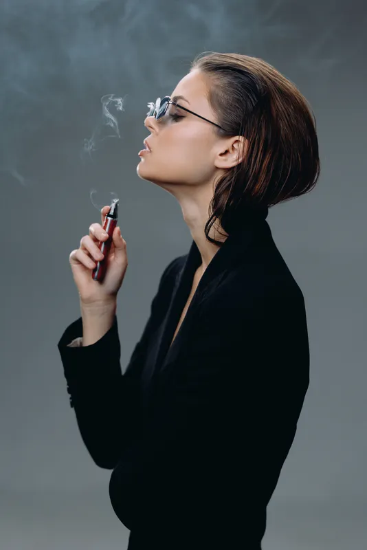 Stylish woman smoking electronic cigarette with smoke on grey background, wearing black outfit and sunglasses