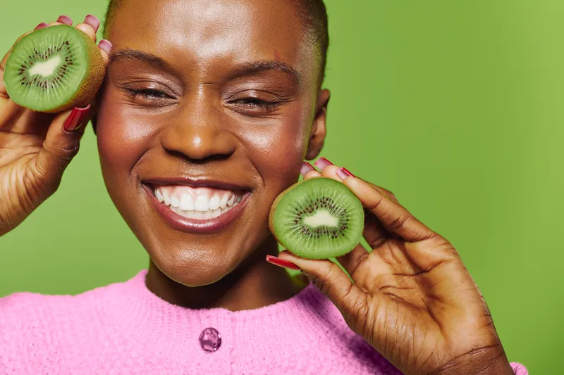 Happy African woman with dark skin holding two kiwi halves near face smiles with closed eyes on green background. Concept of healthy food, fruit, wellness, and natural beauty.
