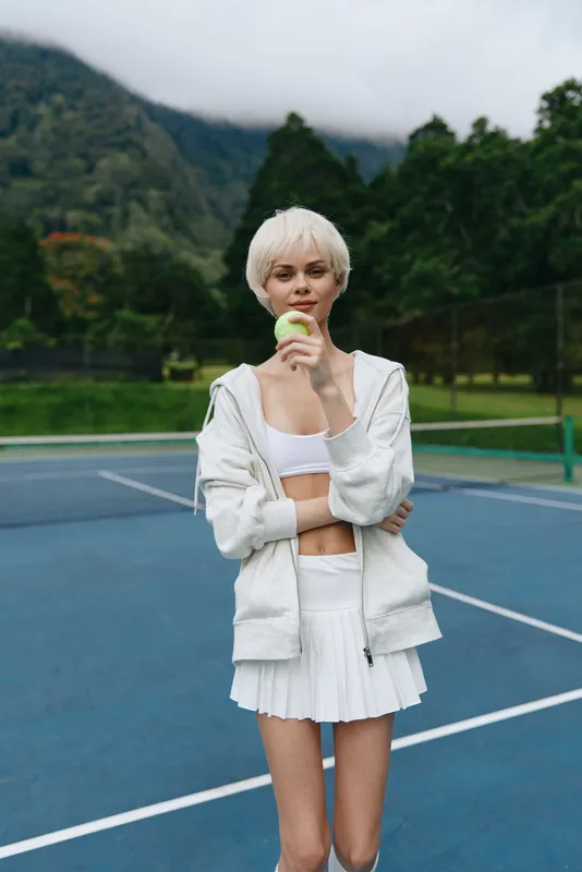 Young woman with short blonde hair, wearing a white sports outfit, holding a green tennis ball on a blue tennis court with mountains in the background