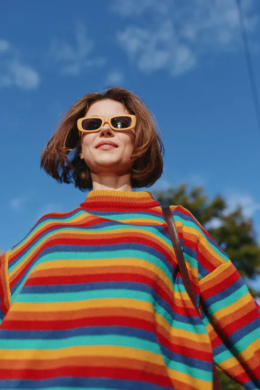 Woman in a colorful striped sweater and yellow sunglasses smiles confidently outdoors against a blue sky. Young adult with bob haircut and casual outfit, relaxed travel lifestyle portrait.
