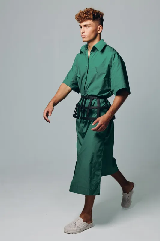 Young man walking in green oversized shirt and matching wide pants with black belt accessory and white slip-on shoes on plain background in studio.