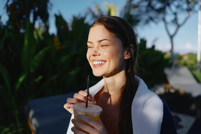 Happy young woman with wet hair enjoys fresh juice outdoors in sunny garden, wrapped in towel, smiling with closed eyes, lifestyle concept with natural light and relaxed mood.
