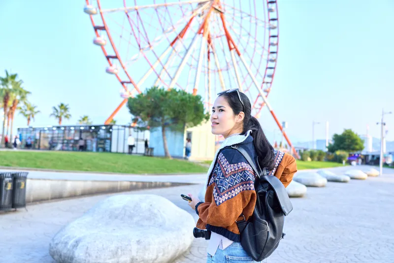 Woman with backpack and camera gazes toward a ferris wheel in a sunny boardwalk park, capturing a travel moment and enjoying outdoor leisure amid bright scenery