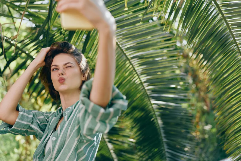 Woman taking a selfie outdoors beneath palm fronds, puckered lips, striped shirt, bright sunny mood, casual summer moment captured in nature.