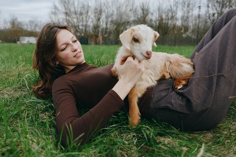 Young woman lies on grass holding a baby goat on her lap in a natural outdoor setting during autumn, enjoying a peaceful moment with the animal in casual clothing.