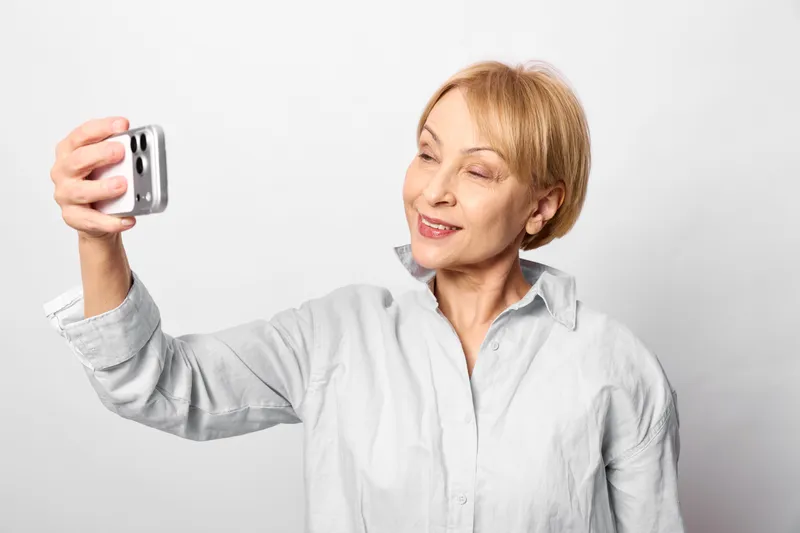Senior woman smiling takes selfie with smartphone, portrait in studio, technology and communication theme, casual shirt, relaxed hand gesture, short hair, light neutral background