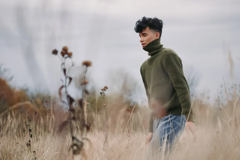 Young man in a green knit turtleneck walks through tall dry grass, captured in an authentic, realistic natural light portrait that highlights quiet introspection and genuine presence. (160 characters)