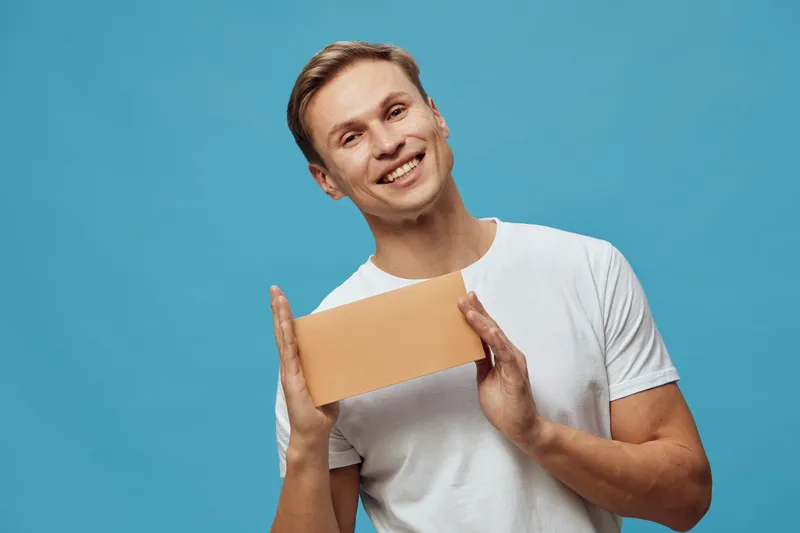 Smiling man holding blank cardboard sign, casual white T-shirt, expressive and cheerful, studio portrait with plain blue background, lifestyle and advertising concept.