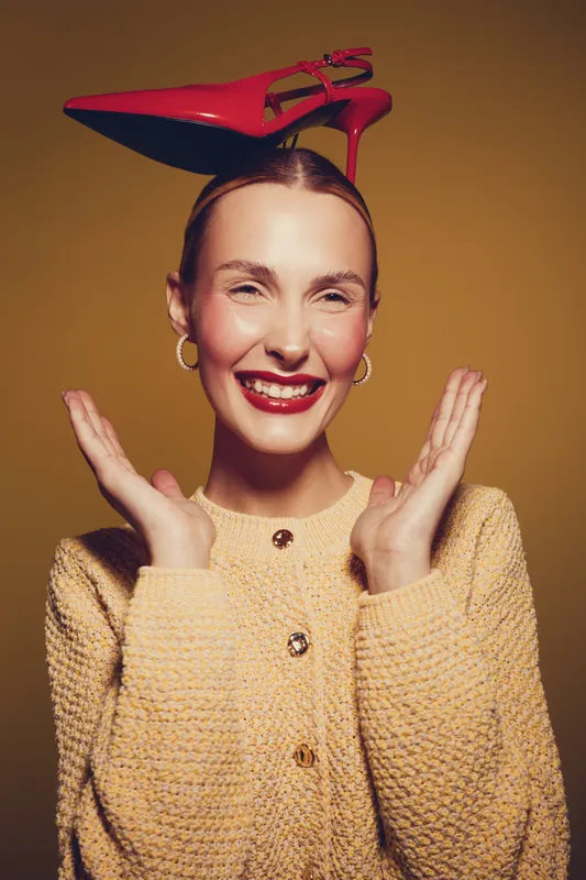 Smiling woman with red high heel shoe balanced on her head wears yellow knitted sweater and gold hoop earrings. Studio portrait with warm lighting and playful fashion concept.