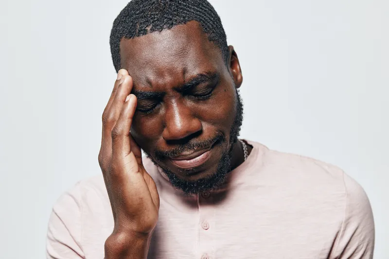 African man with dark skin holding head with hand, showing pain or headache, wearing light pink shirt on white background. Male portrait with closed eyes and stressed expression.