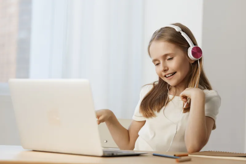 Cheerful girl enjoying music at home, wearing headphones, sitting at a laptop with a bright background, conveying a sense of joy and creativity