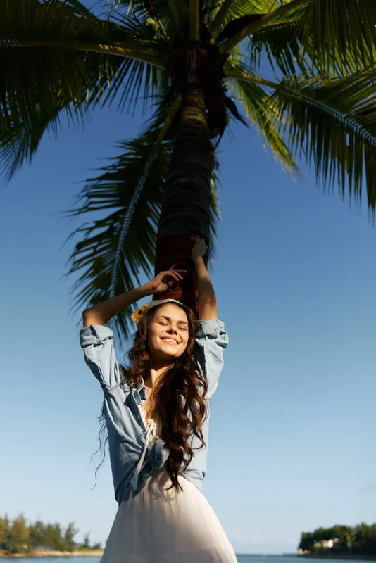 Smiling woman enjoying tropical nature under a palm tree with clear blue sky and sunlight, expressing happiness and freedom outdoors.