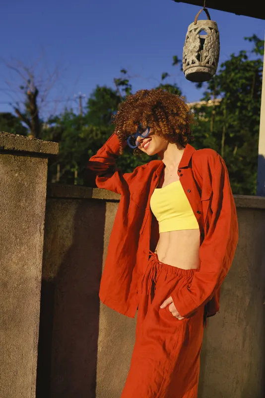 Young woman with curly hair wears a red suit and yellow crop top, posing outdoors in bright sunlight with a bold street style against a stone wall and a hanging lantern overhead