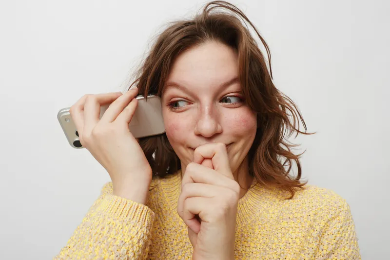 Young woman in yellow sweater holds smartphone to ear, smiling shyly with hand near mouth. White background, casual style, concept of communication, happiness, and social connection.