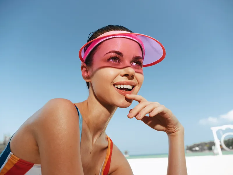 Bright portrait of a joyful woman at the beach wearing a pink visor, sunlit skin, and a colorful swimsuit, captured under a clear blue sky and warm sunlight.