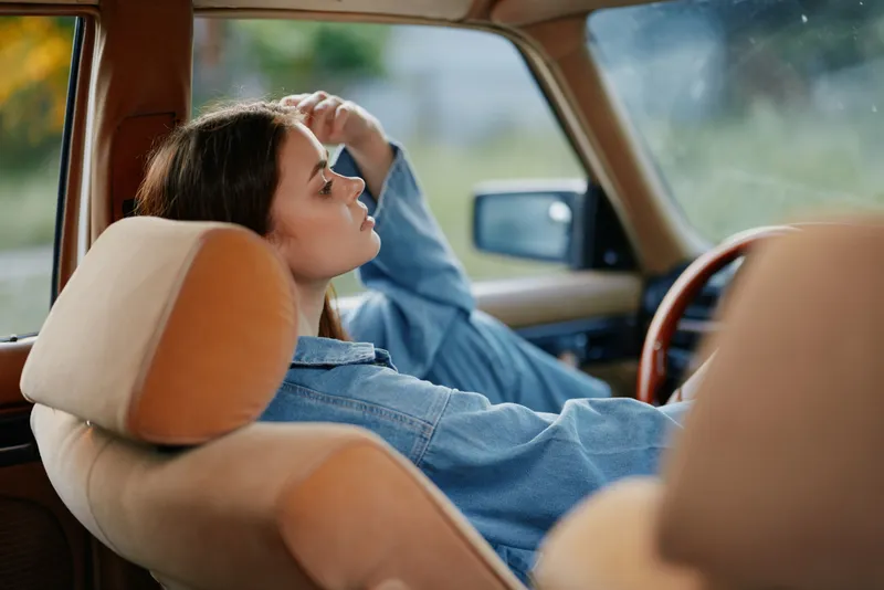 Thoughtful young woman in a denim jacket relaxing inside a vintage car, with serene emotions and soft natural light setting a calm mood.