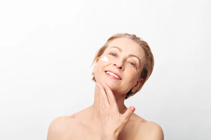 Mature woman with short hair gently touches her face with hand smiling softly on white background. Concept of natural beauty, skincare, self-care, aging gracefully and wellness.