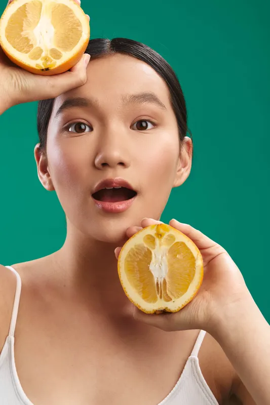 Asian woman playfully holds orange halves against a bright green background, smiling with curiosity. The fresh citrus scene conveys energy, vitality, and a healthy lifestyle in a simple studio