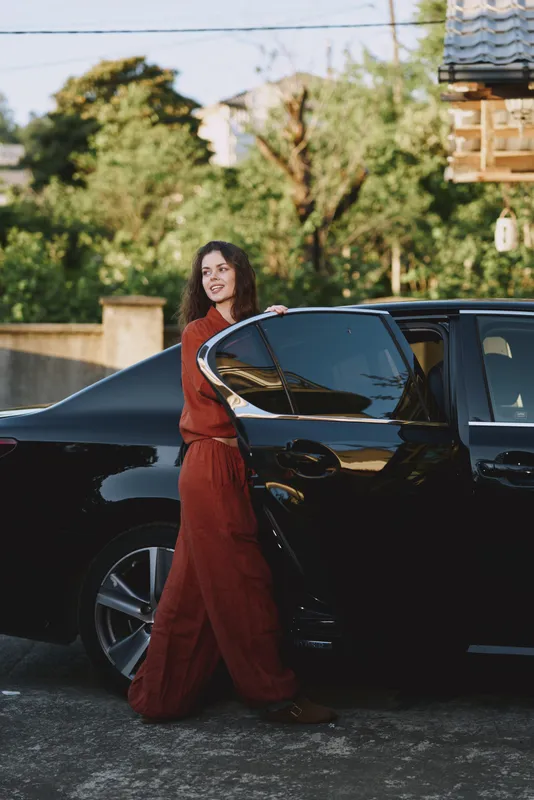 A stylish woman in a red dress stands beside a sleek black car in a sunlit driveway posing confidently for a lifestyle portrait, urban vibe, natural light, candid moment, fashionable pose