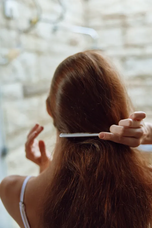Woman combing long brown hair in soft natural light, close-up rear view with blurred stone background creating calm and intimate atmosphere.
