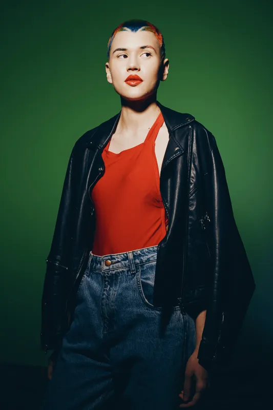 Young woman with short colorful hair wearing a red top and leather jacket, standing confidently against a green background in casual denim jeans.