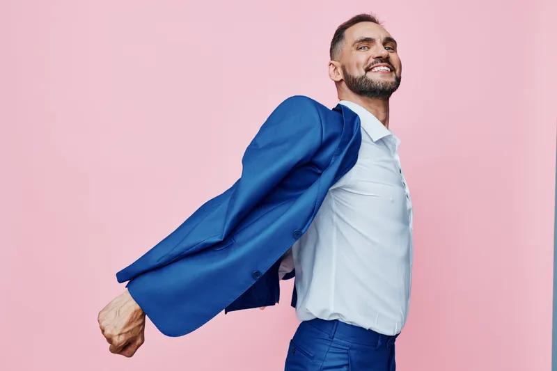 Professional man in business attire with a blazer off, standing against a solid pink background, expressing confidence, energy, and pride in a dynamic pose.