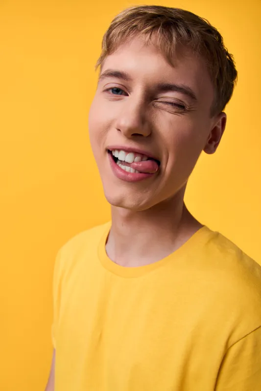 Young man wearing a yellow shirt winking and sticking out his tongue against a bright yellow background, playful expression, casual vibe, lighthearted energy, vibrant mood